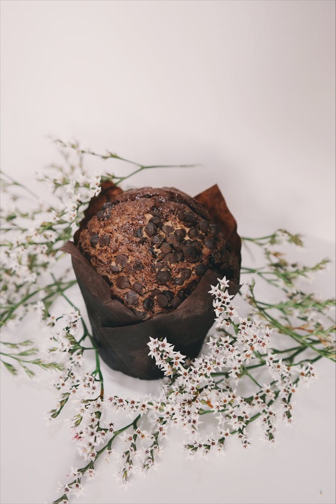 Chocolate muffin surrounded by white flowers on a neutral background.