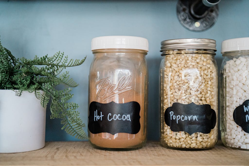 Glass jars with popcorn, hot cocoa, and marshmallows on a kitchen shelf with a potted plant.