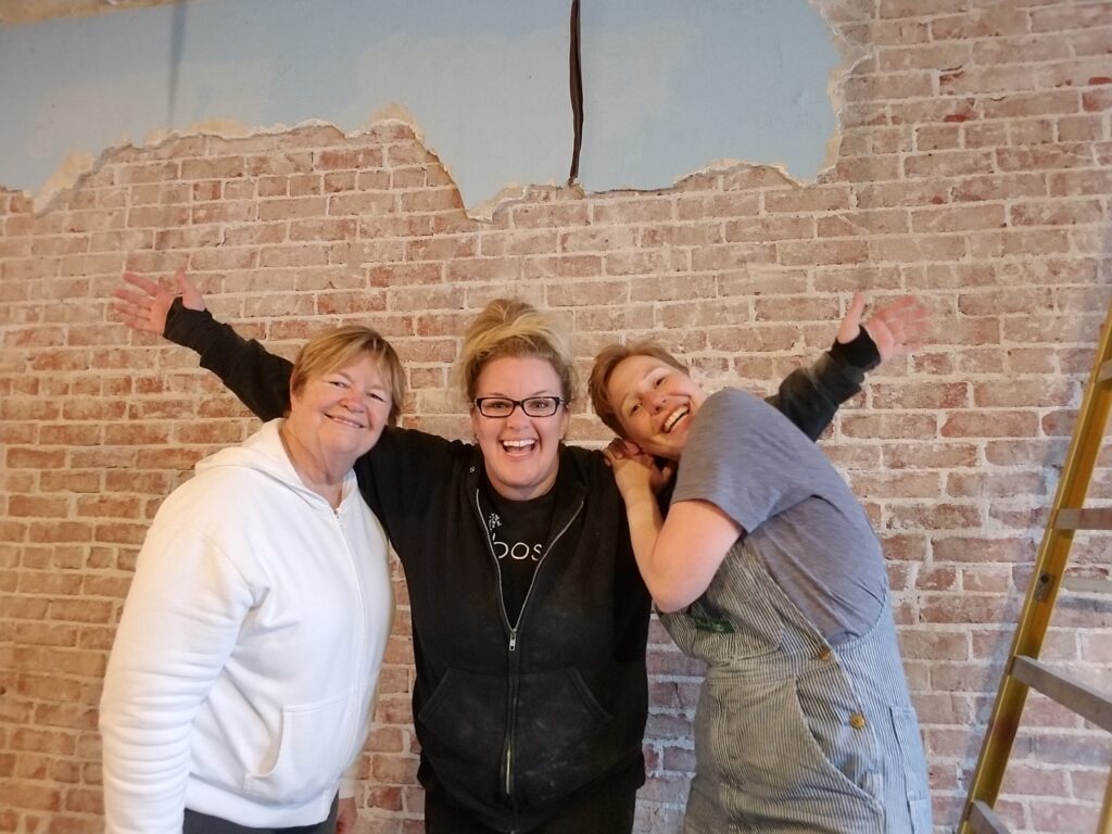 three women stand proudly in front of a antique brick wall that they exposed. 