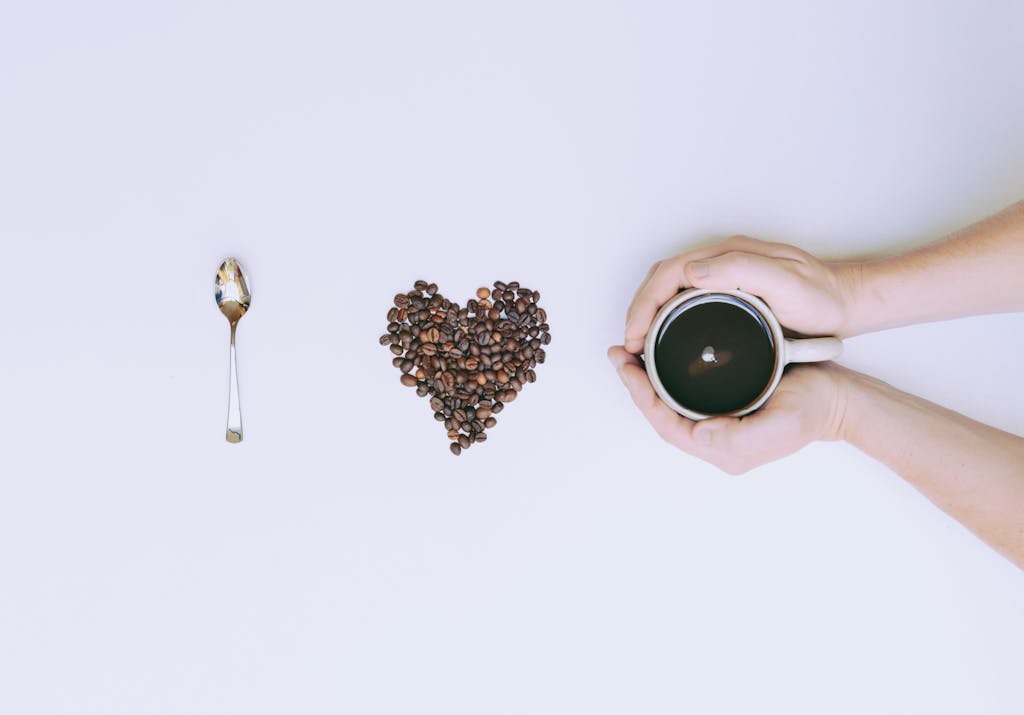 Coffee beans arranged in a heart shape with a mug and spoon on a white background.