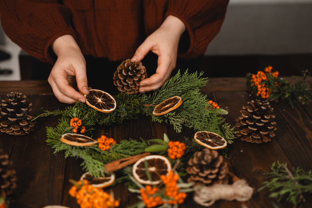 Hands arranging pine cones and dried oranges on a Christmas wreath.