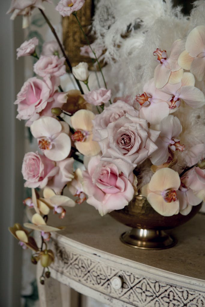 Vintage metal vase with composition of orchid and rose and pink flowers with feathers placed on figured wooden cabinet in vintage style room