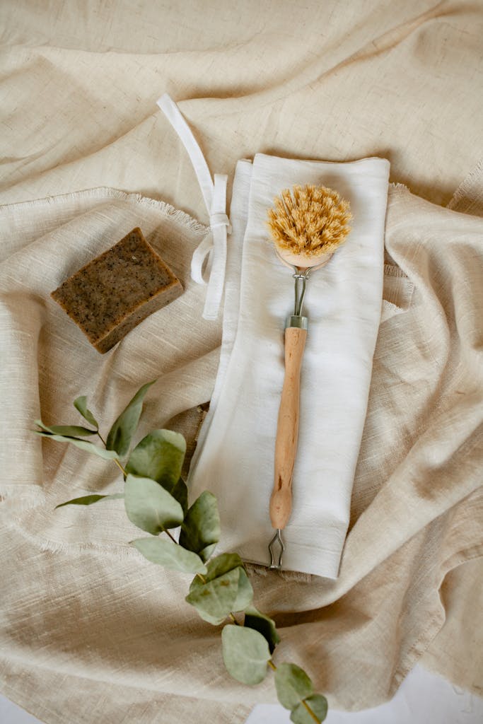 Flat lay of eco-friendly cleaning supplies, including a wooden brush and linen cloth, on soft beige fabric.