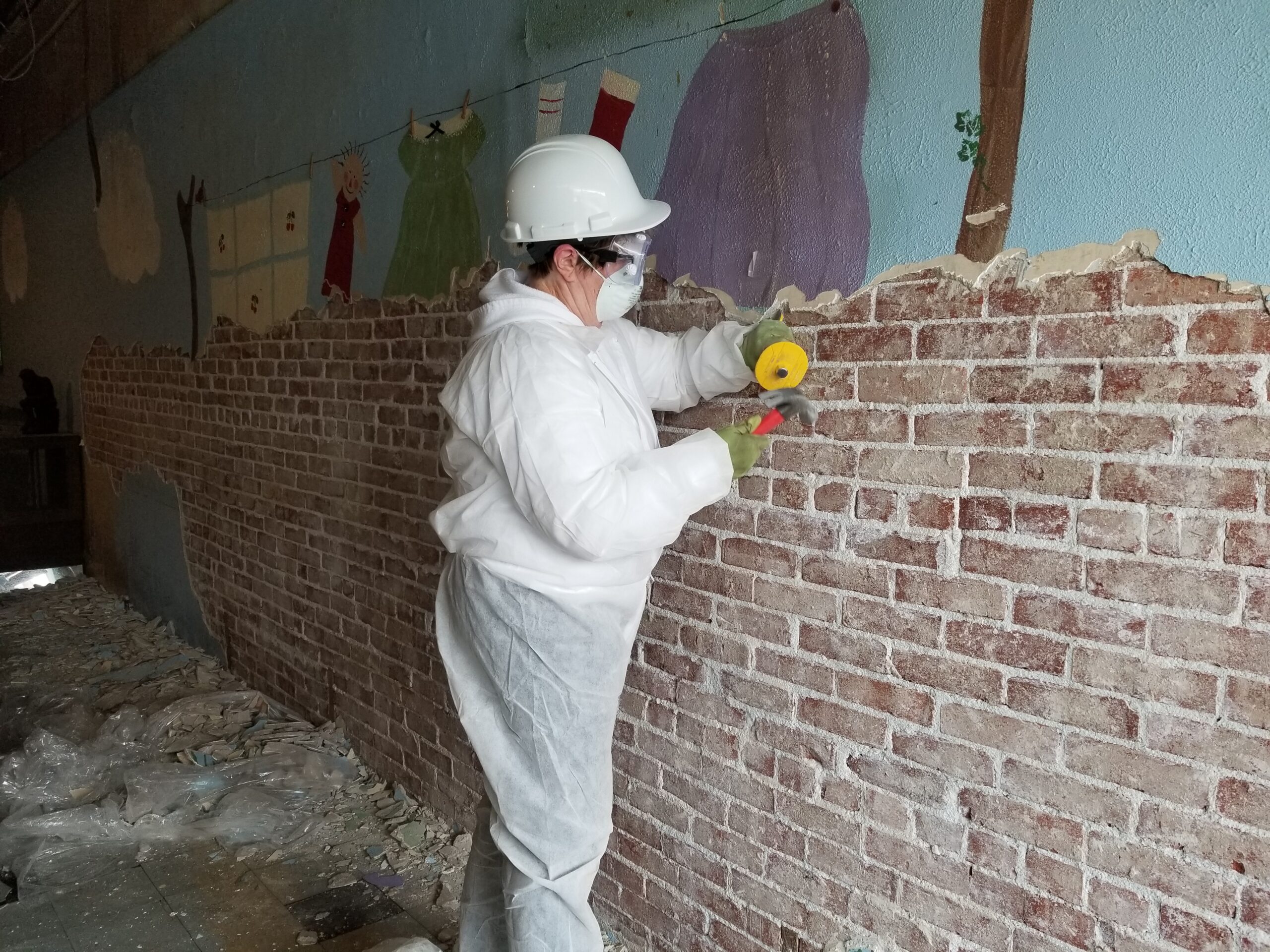 A woman dressed in protective clothing using a chisel to remove plaster from an antique brick wall