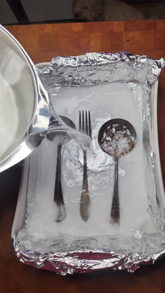 Boiling water is poured into a casserole dish with antique silverware,  baking soda,  and aluminum foil 