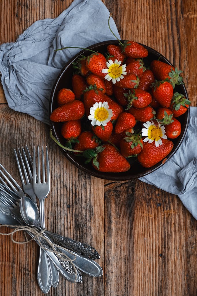 A bowl of fresh strawberries with daisies on a rustic wooden table, surrounded by silverware.