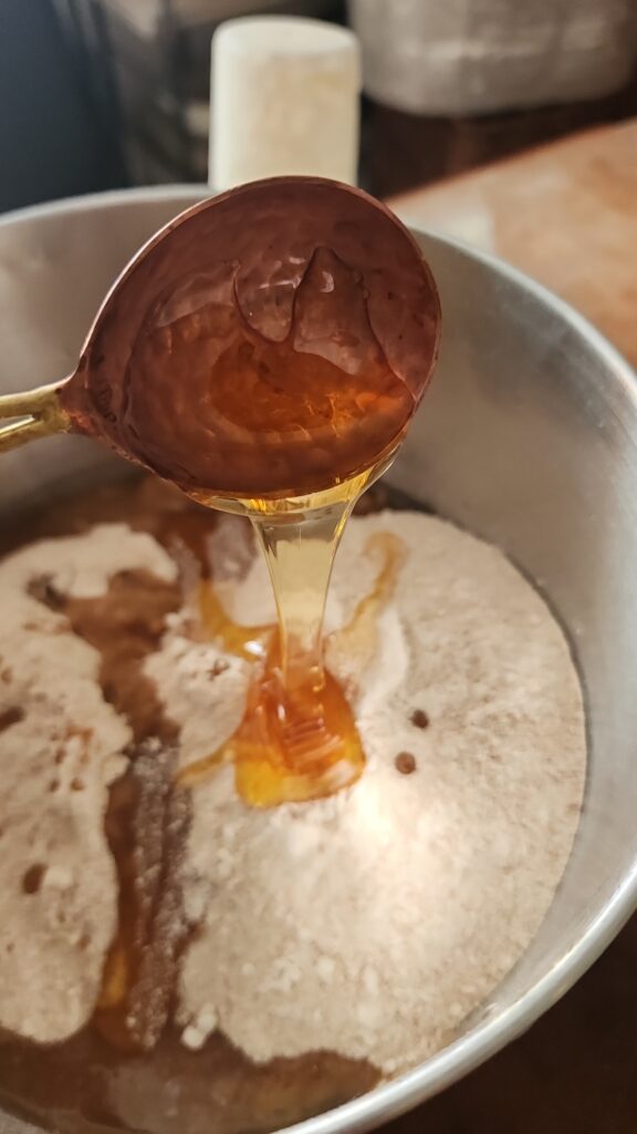 Raw honey is being poured into a mixing bowl of bread ingredients to make whole grain sandwich bread.