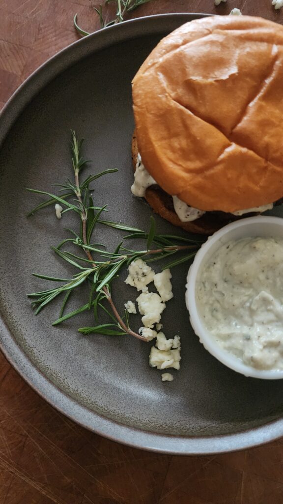 Delicious made-from-scratch blue cheese & rosemary mayo gently oozes out of a freshly cooked burger with brioche bun. It is sitting on a gray plate that is garnished with a fresh rosemary sprig, blue cheese crumbles, and a small bowl of blue cheese rosemary mayo on the side.