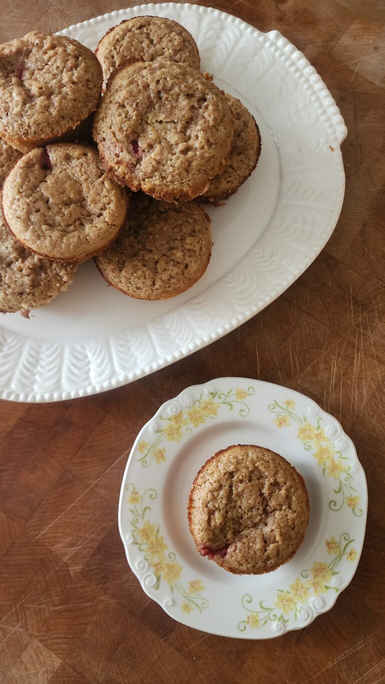 A vintage white platter filled with golden-brown sourdough raspberry chocolate chip muffins, alongside a single muffin served on a floral antique dessert plate, all set on a rustic wooden surface.