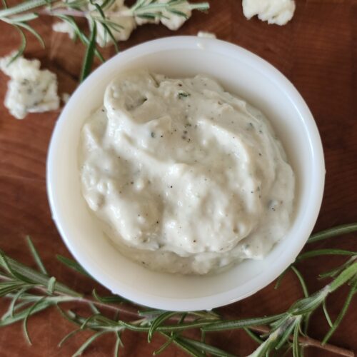 Freshly made rosemary and blue cheese sauce is ready to eat in a beautiful antique white dish on a wooden surface. Sprigs of fresh rosemary and blue cheese crumbles are strategically placed around the dish.