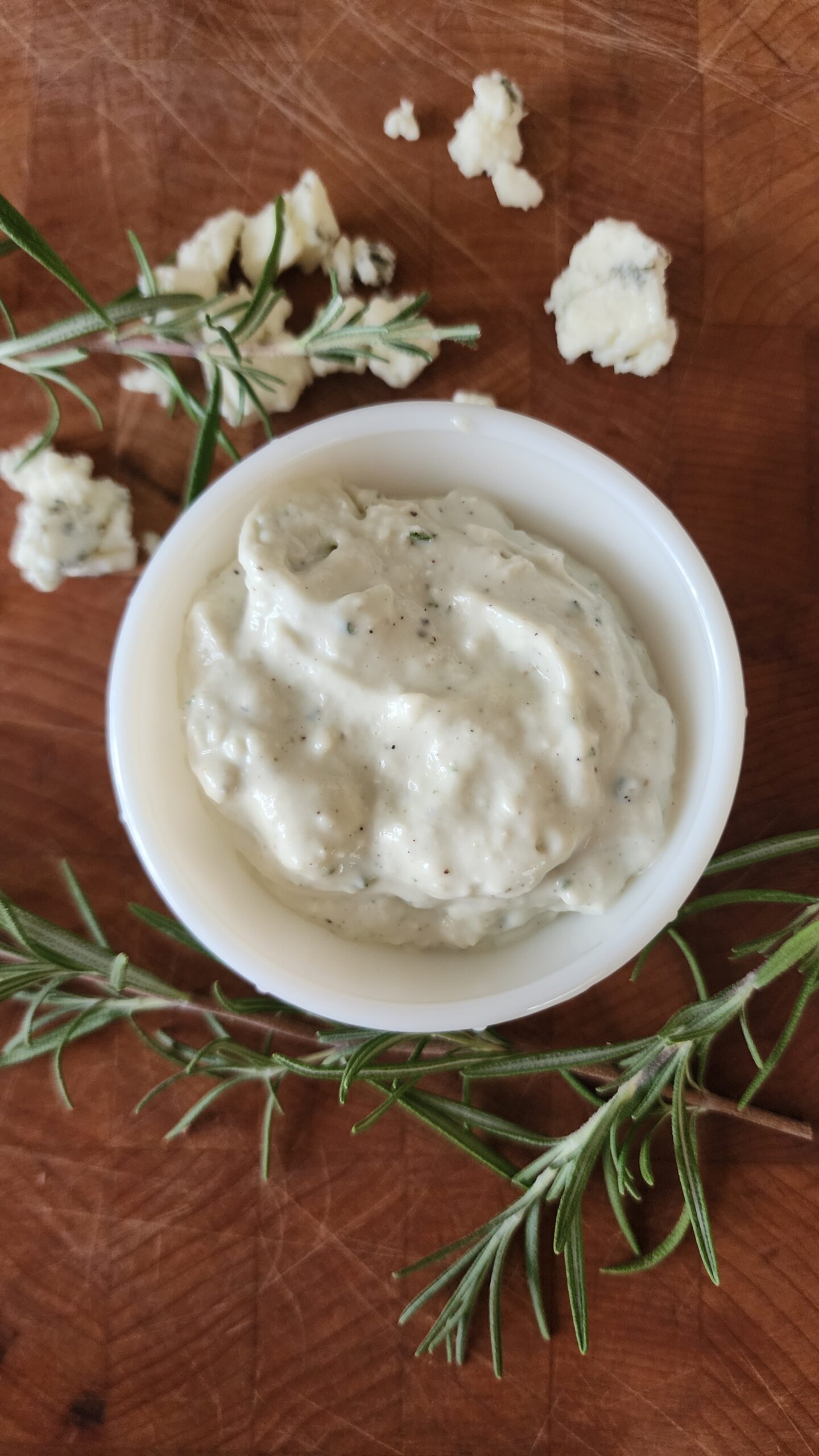 Freshly made rosemary and blue cheese sauce is ready to eat in a beautiful antique white dish on a wooden surface. Sprigs of fresh rosemary and blue cheese crumbles are strategically placed around the dish.