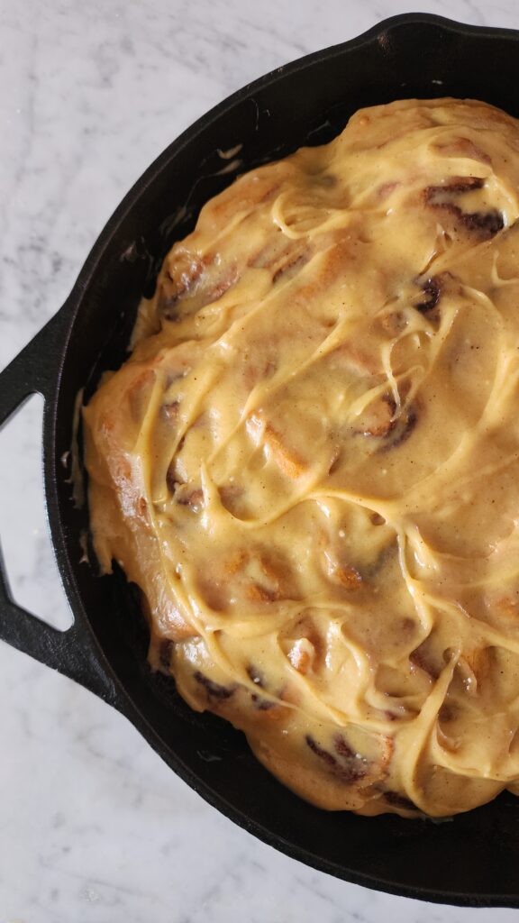 Freshly iced Sourdough Pumpkin Cinnamon Rolls in a cast iron pan on a marble surface