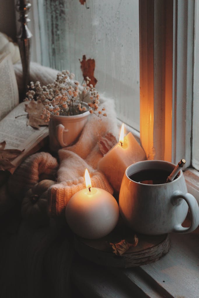 A warm, cozy setting featuring a cup of tea, candlelight, and autumn decor near a rainy window.