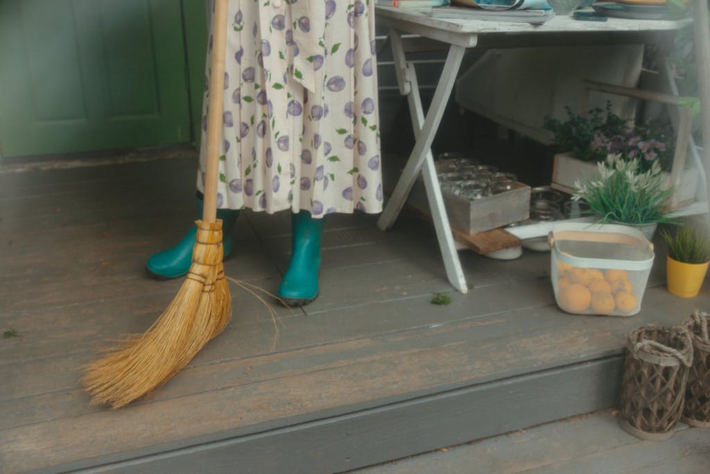 A woman in a floral dress and rubber boots sweeps a wooden porch beside a table with plants.