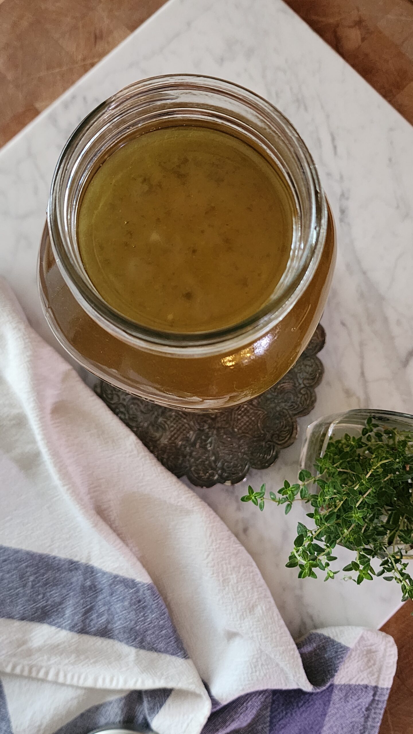 Freshly made chicken bone broth in a jar surrounded by a tea towel and fresh herbs is ready to enjoy as a gut-healthy beverage or soup base.