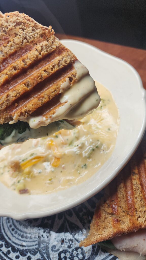 A warm pressed sandwich is being dunked into a bowl of comforting broccoli cheddar soup for the perfect fall meal.