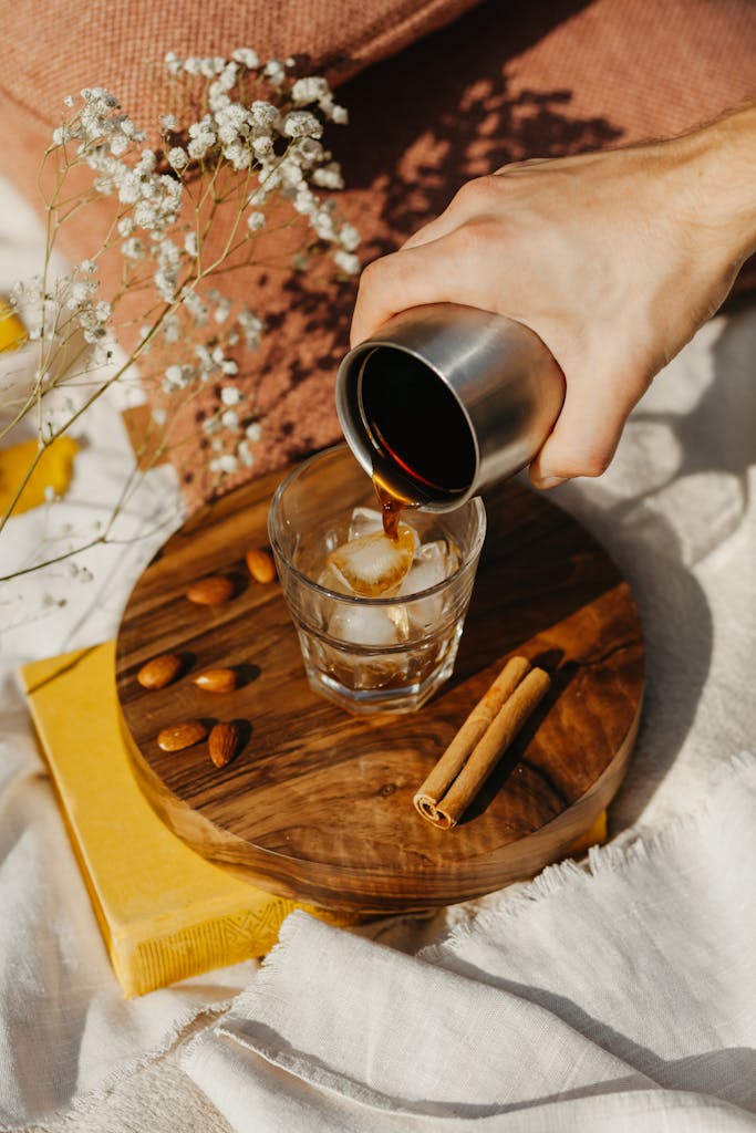Aesthetic coffee pouring scene with glass, ice, almonds, cinnamon, and rustic decor.