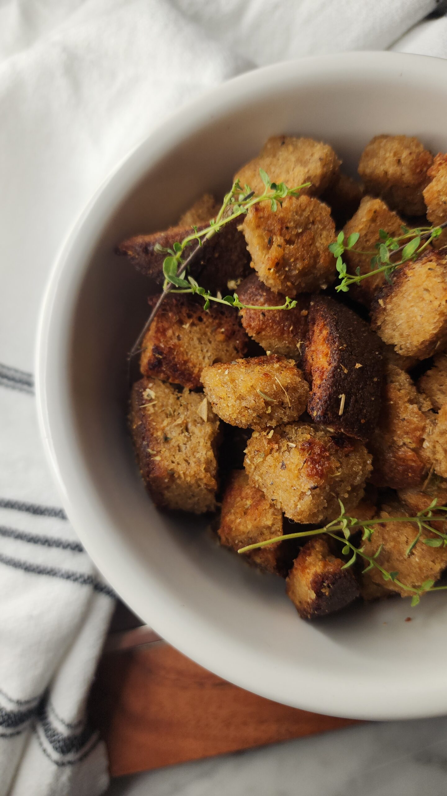 A white bowl is filled with freshly baked golden crunch sourdough croutons with fresh herbs on a wooden surface.