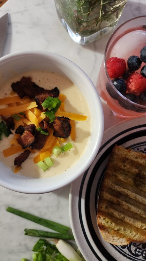A delicious display of creamy chicken bacon ranch soup in a white bowl with a warm pressed sandwich and a glass of bubbly and fruity kefir water.