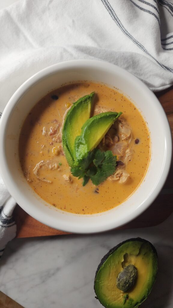 Bowl of creamy taco soup topped with sliced avocado and fresh cilantro, served in a white bowl on a marble surface with a striped towel.