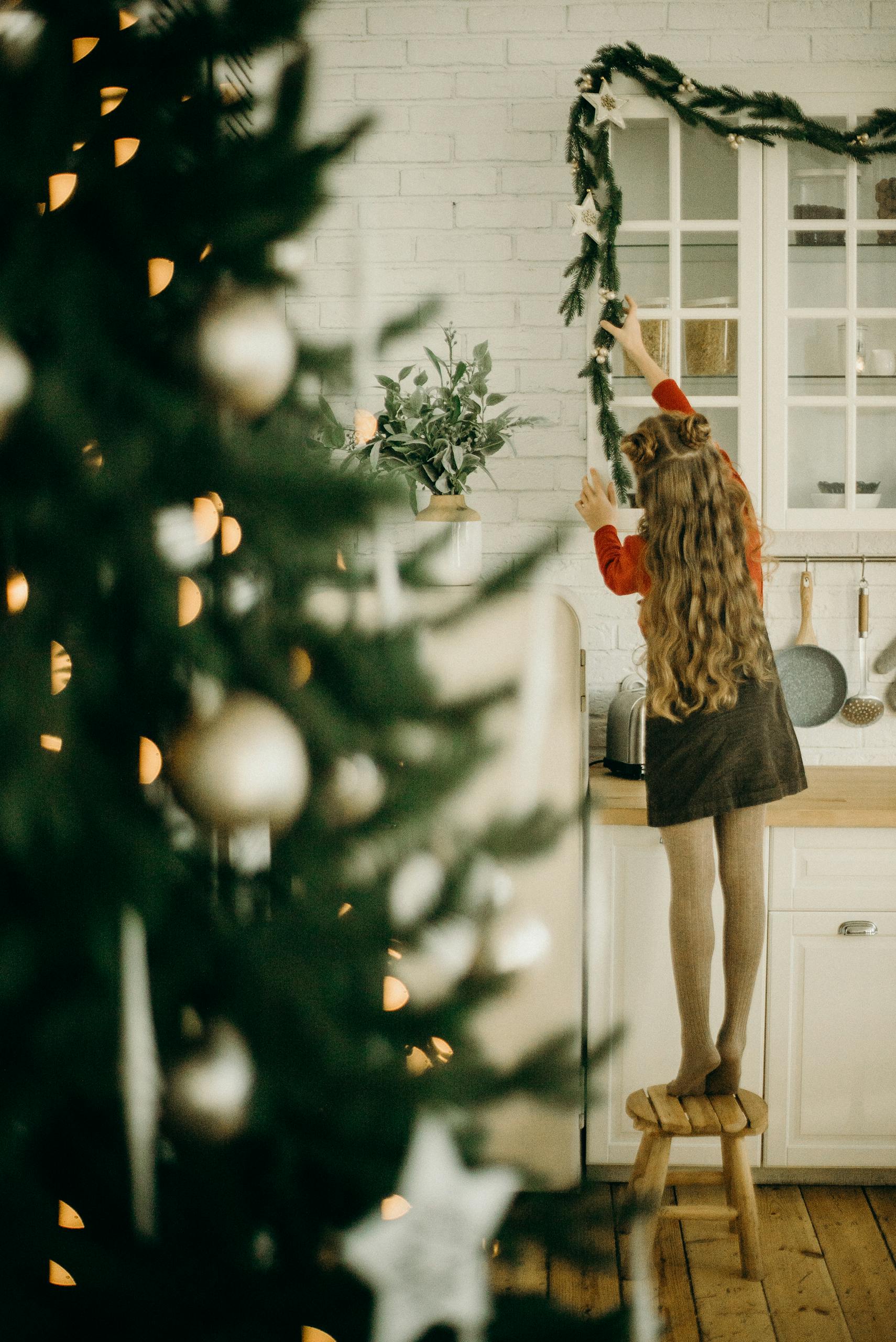 A young girl stands on a stool decorating a kitchen with festive Christmas garlands.