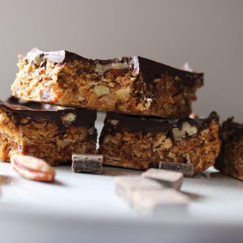 Healthy Pecan Date Bars are stacked thoughtfully and displayed on a white plate surrounded by dark chocolate chunks and pecans.