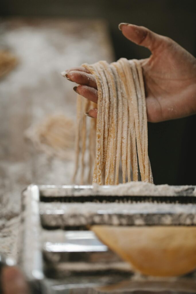 Close-up of a hand pulling fresh pasta from a pasta cutter, dusted with flour, in a kitchen setting.