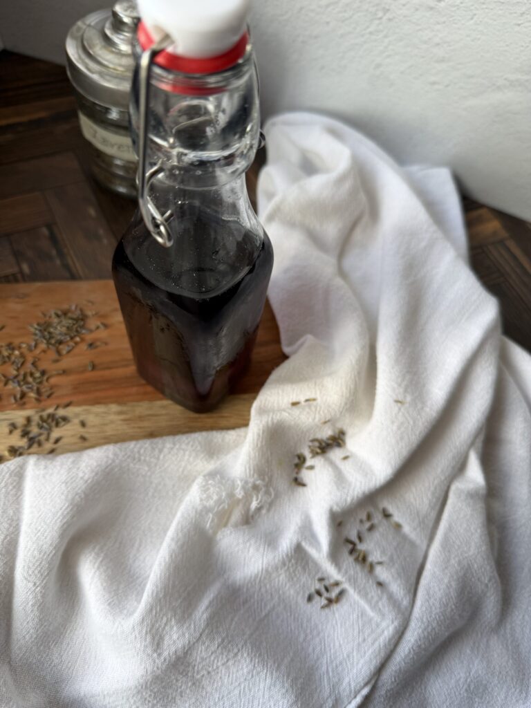 A lavender simple syrup in a bottle on a wooden surface with a tea towel and fresh lavender buds. 