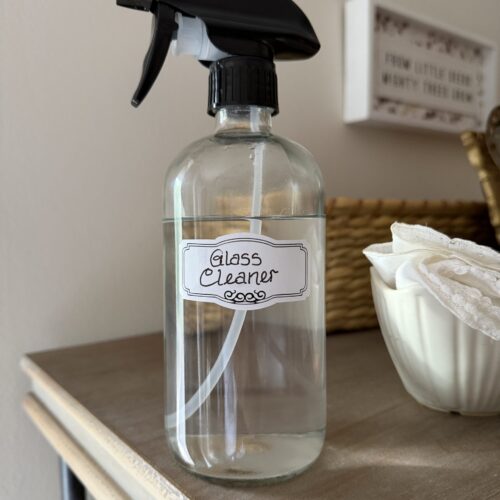 Homemade glass cleaner in a labeled clear glass spray bottle on a wooden surface.