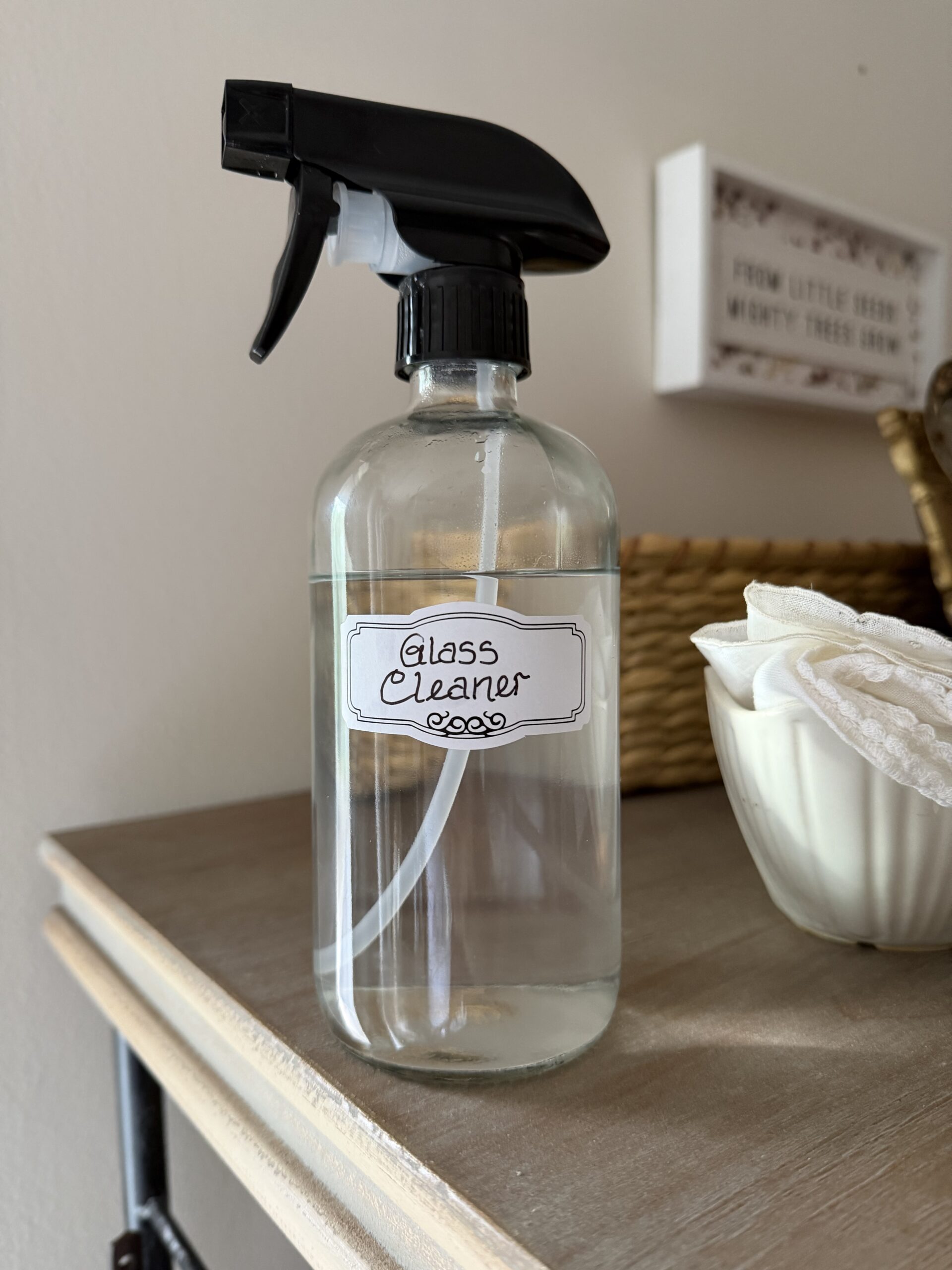 Homemade glass cleaner in a labeled clear glass spray bottle on a wooden surface.