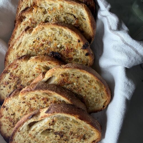 A golden stack of buttery sourdough garlic toast in a tea towel lined bread basket.