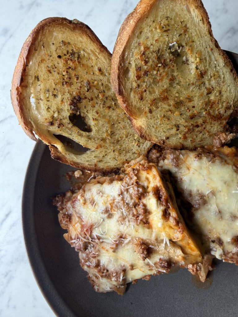 Two slices of sourdough garlic toast on a plate with slices of cheesy lasagna made with sourdough noodles.