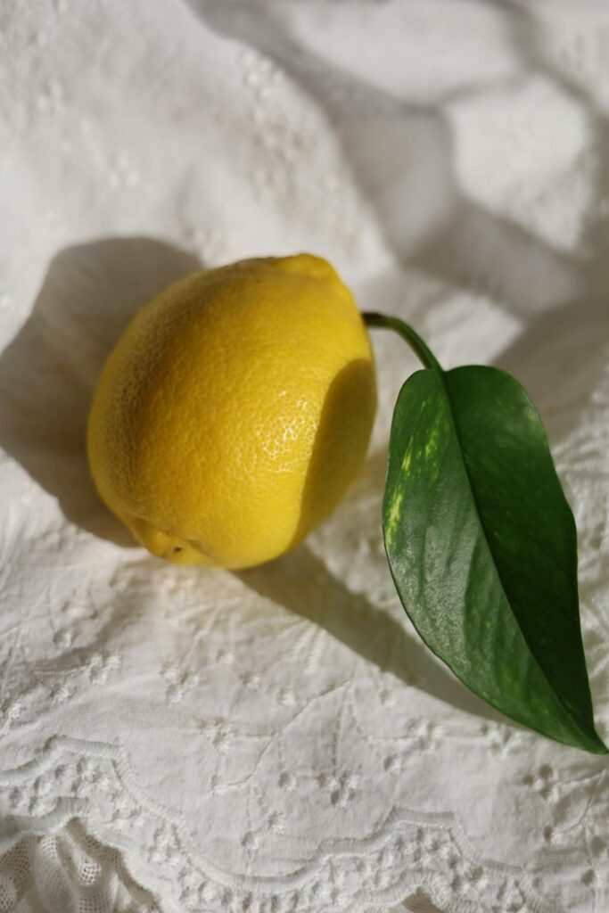 A fresh lemon with a green leaf on a white textured fabric surface.