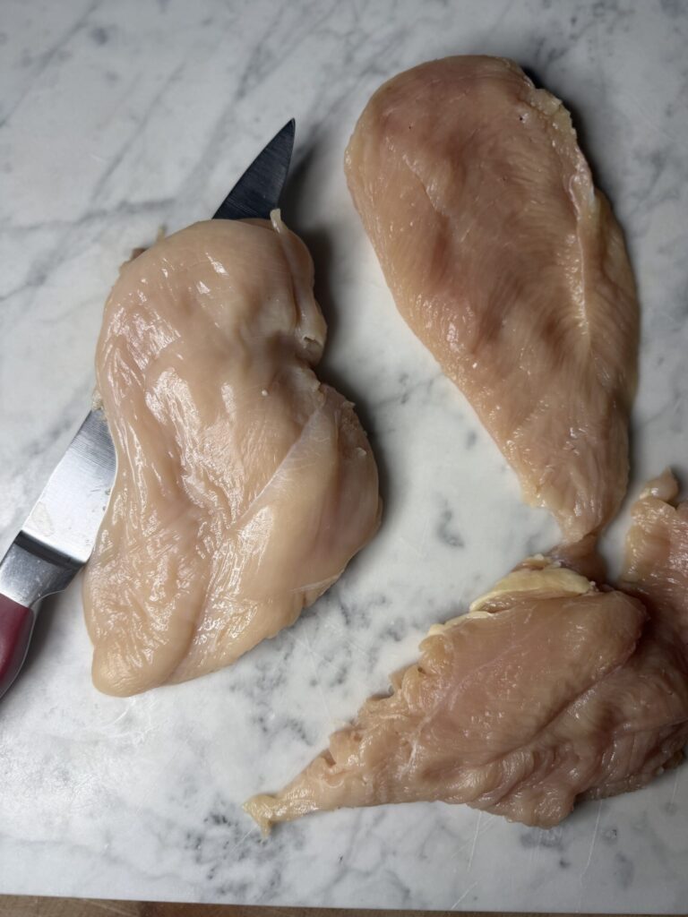 A knife is fileting a chicken breast on a marble surface. 