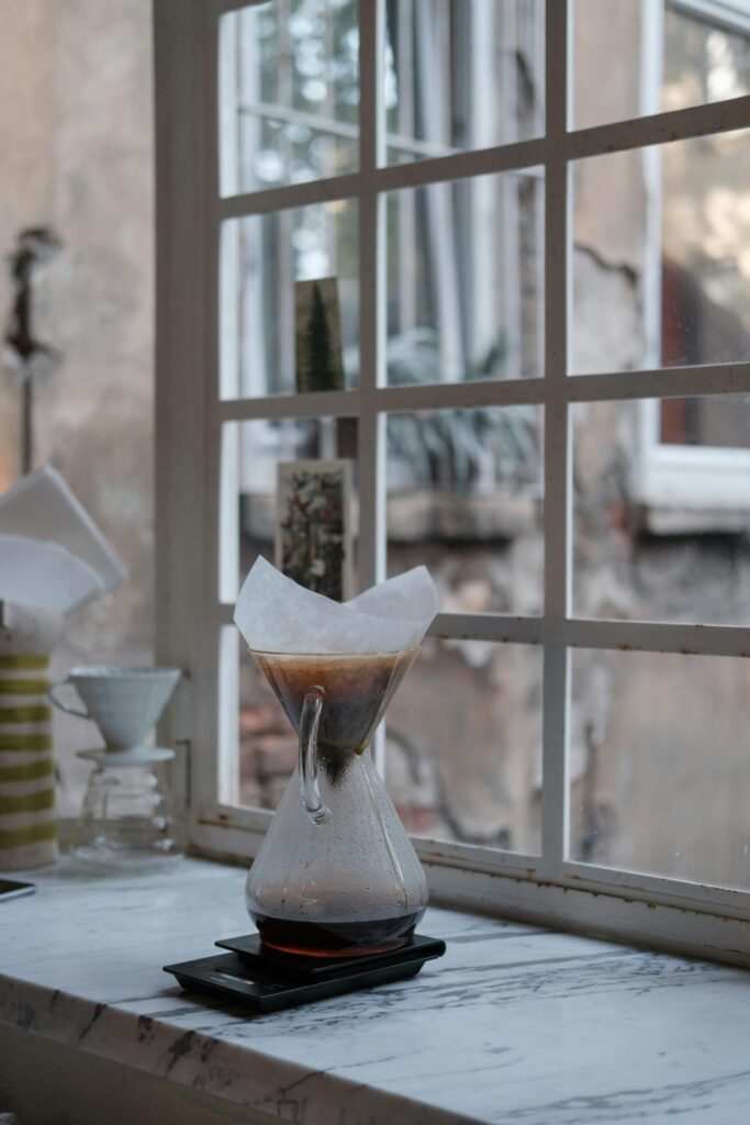 A drip coffee maker with brewed coffee in a homey setting by the window.