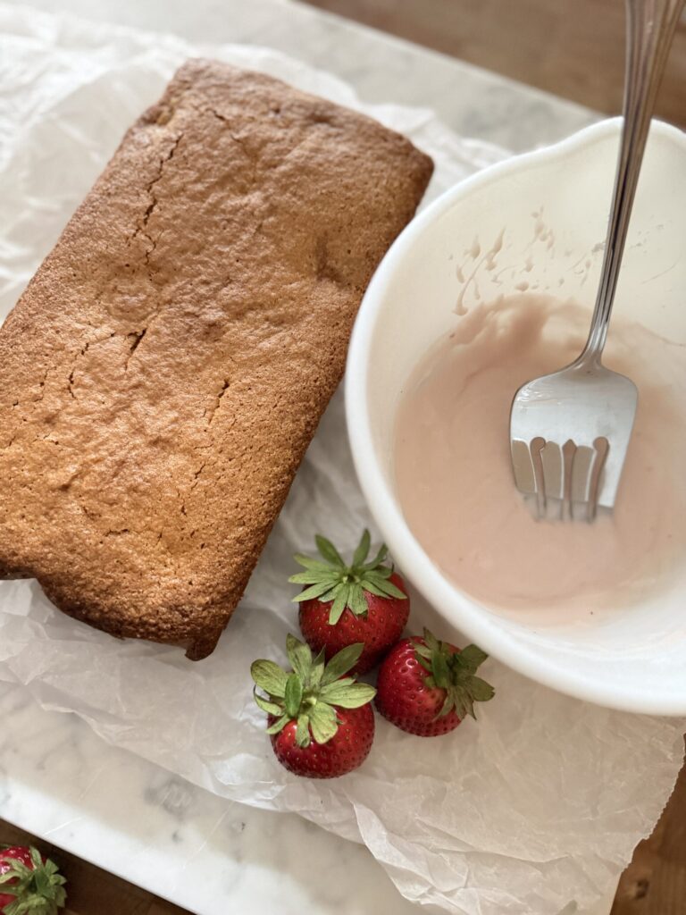 A baked loaf of sourdough strawberry quick bread on parchment paper with a bowl of strawberry icing, ready to be glazed. 