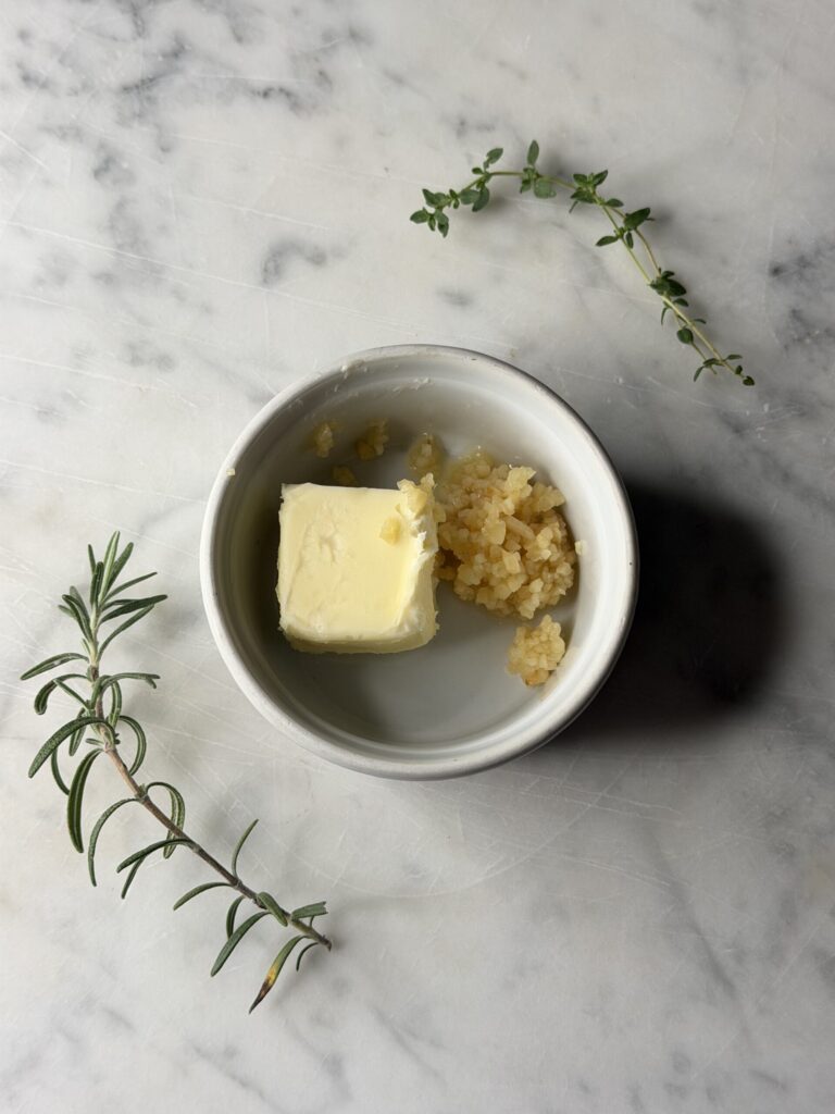 Butter and minced garlic in a white ramekin surrounded by fresh herbs on a marble surface. 