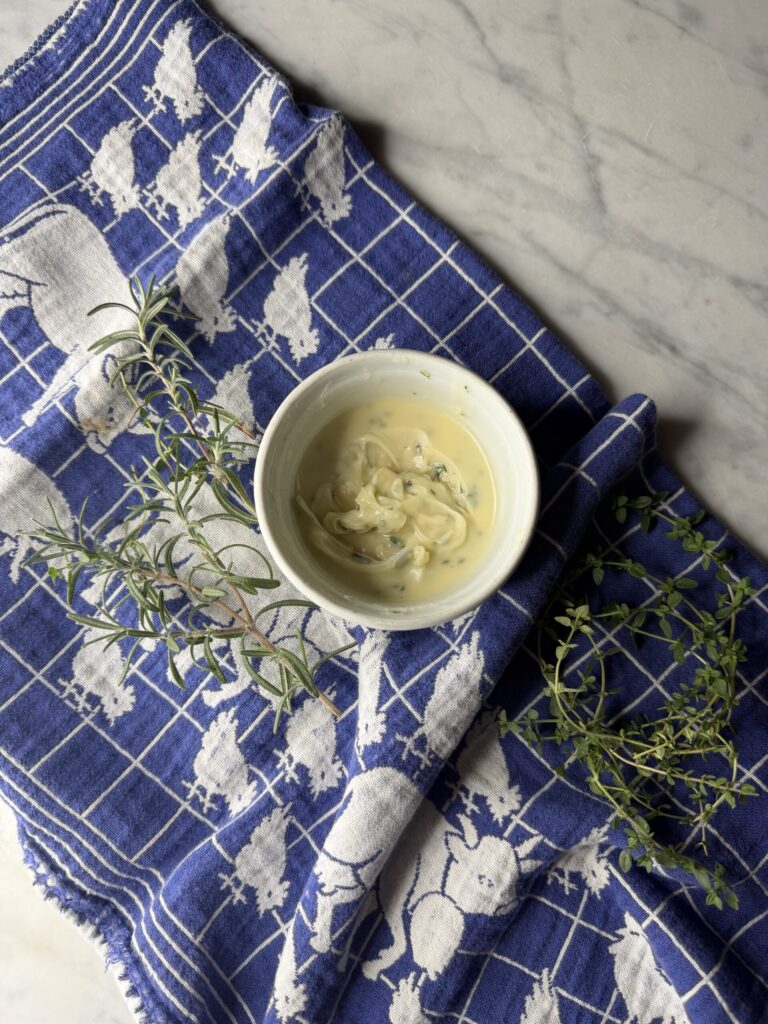 Creamy herbed compound butter in a small white dish on a vintage dish towel. 