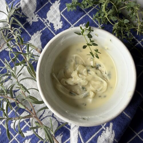 Softened butter with fresh rosemary and thyme in a white bowl on a tea towel.