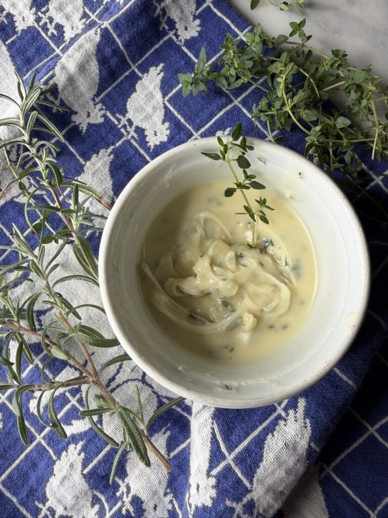 Softened butter with fresh rosemary and thyme in a white bowl on a tea towel.