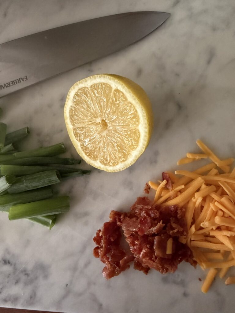 A sliced lemon, green onion, chopped crispy bacon, and shredded cheddar cheese on a marble surface.