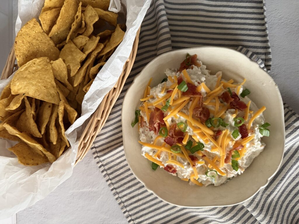 A hefty bowl of creamy bacon ranch dip with a basket full of fresh tortilla chips for serving.