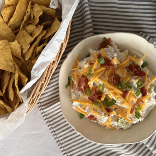A hefty bowl of creamy bacon ranch dip with a basket full of fresh tortilla chips for serving.