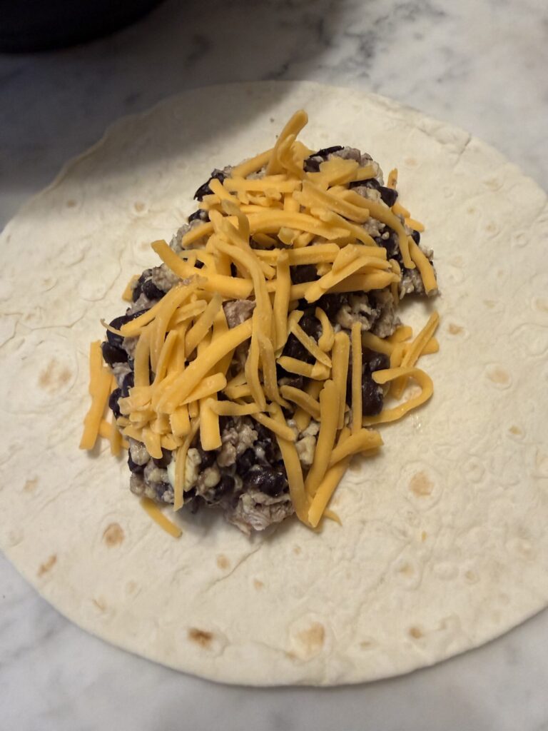 A sourdough tortilla is filled with the breakfast burrito mixture and shredded cheddar cheese before being rolled.