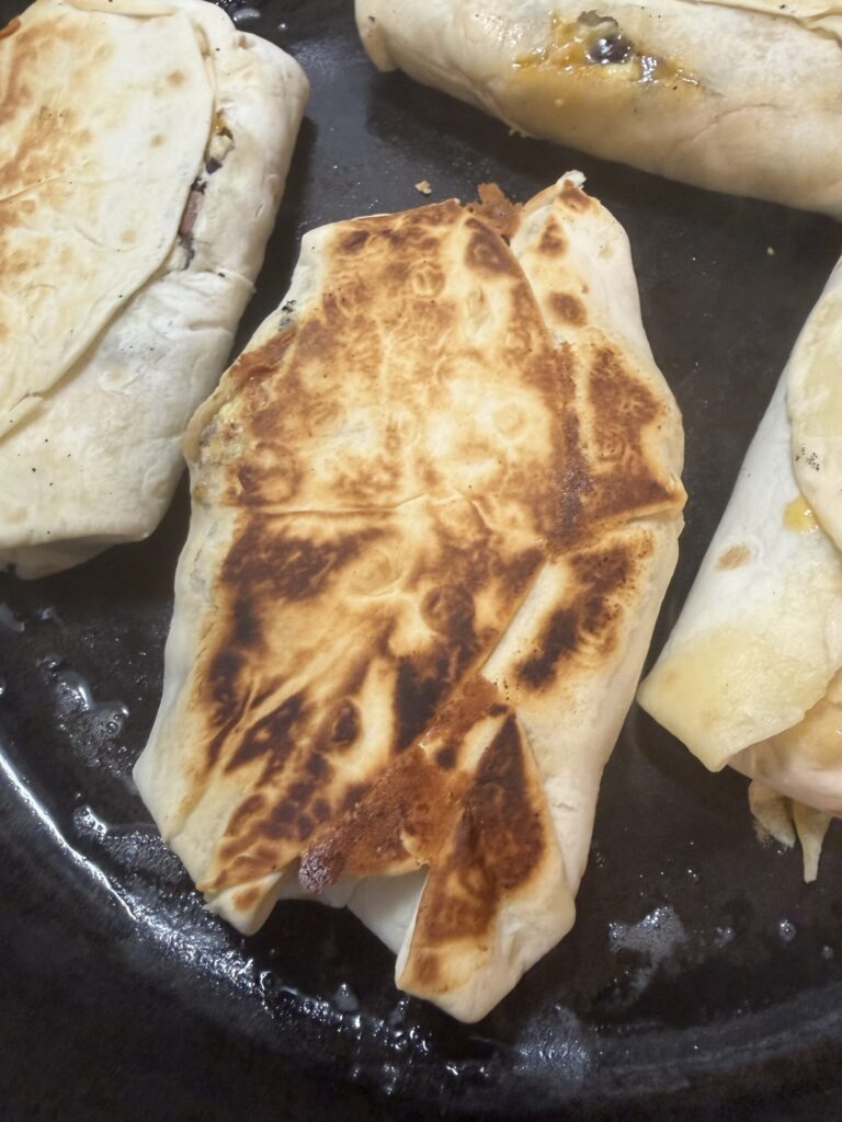 Breakfast burritos are cooking in a cast iron skillet until the tortillas are golden brown.