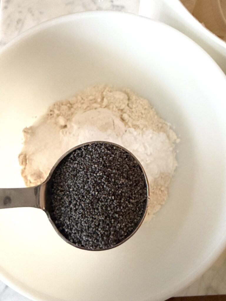 Poppy seeds are added to a bowl of dry ingredients for sourdough lemon muffins.