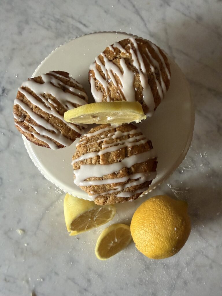Three lemon sourdough muffins with a lemon glaze on a cake stand with sliced lemons on a marble surface.
