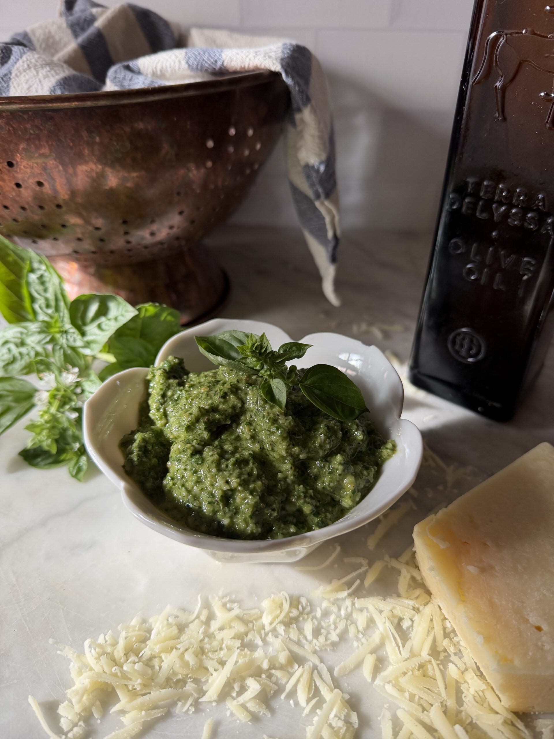 A bowl of freshly made homemade basil walnut pesto on a marble surface with grated parmesan cheese, olive oil, and fresh basil leaves.