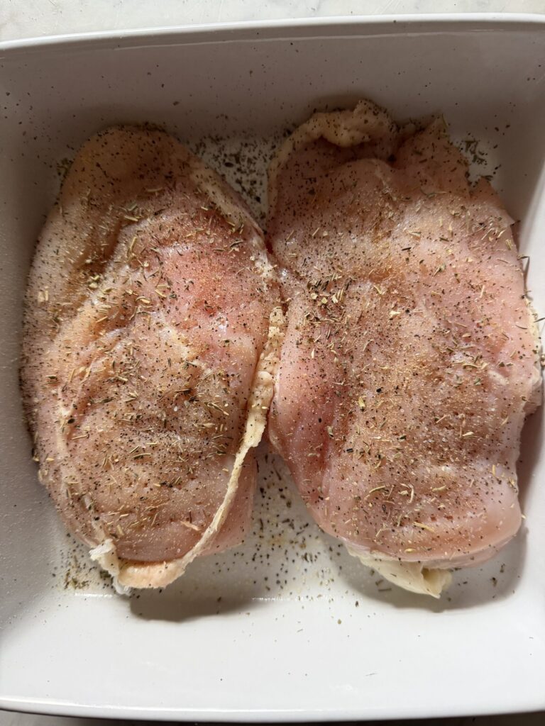 Raw chicken breasts in a baking dish covered in spices and herbs.