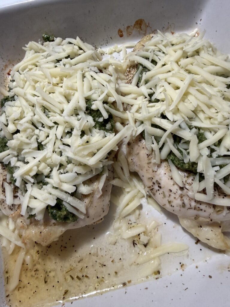 Chicken breast is covered with fresh basil walnut pesto and shredded mozzarella cheese in a baking dish ready to bake.